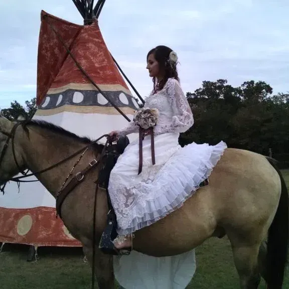 Girl in a white dress riding a brown horse beside a teepee outdoors