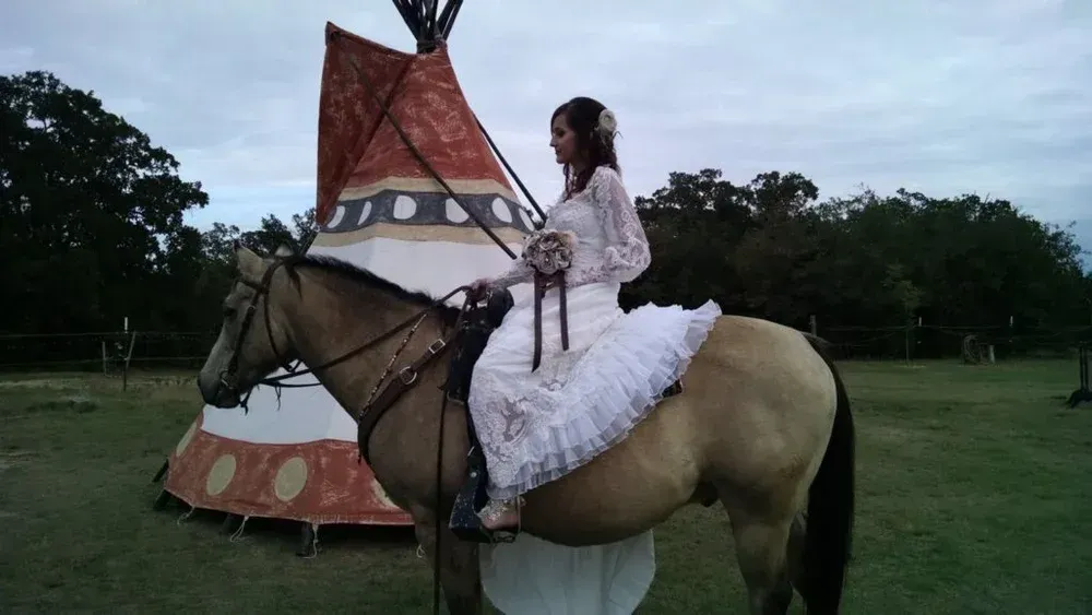 Person in white dress riding a horse beside a painted teepee in a park at dusk