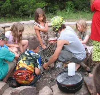 Children sitting around a campfire cooking over a black pot outdoors