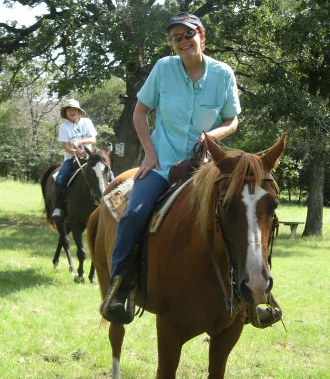 Two people riding horses on a sunny grassy trail, with trees in the background.