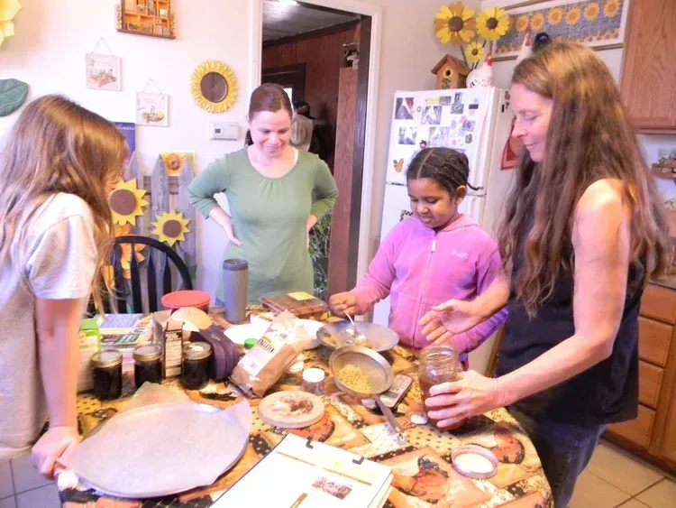 People gathering around a kitchen table, making food together with jars, bowls, and pastries.