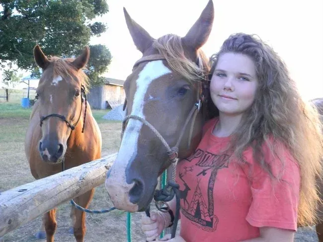 Girl in red shirt standing with two horses by a fence at sunset