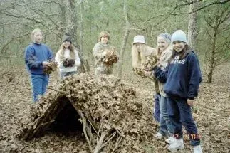 Five people stand outdoors beside a small leaf-and-branch shelter in a wooded area.