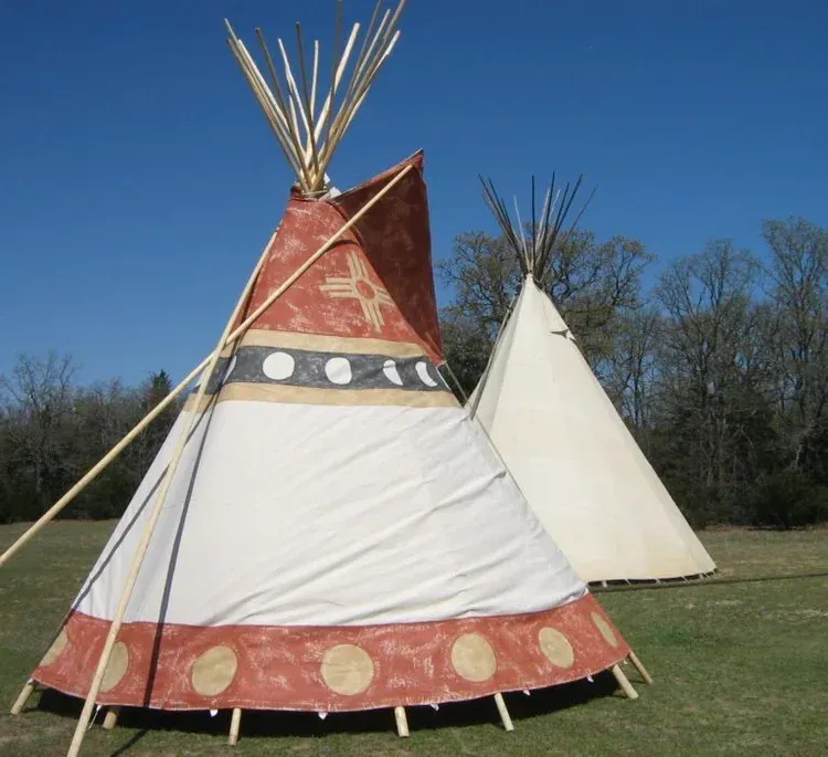 Two teepees on a grassy field, one red-and-white with poles, one plain white, under a blue sky
