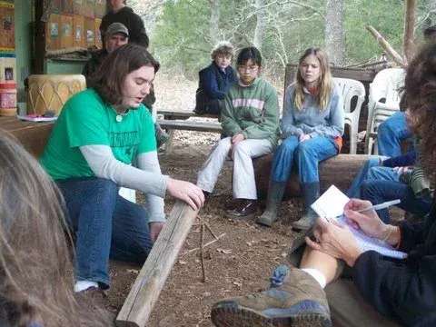 People seated outdoors in a circle during a discussion, with one person speaking on a log bench