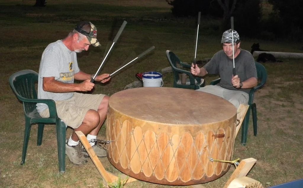 Two people playing drums outdoors at night around a large wooden drum