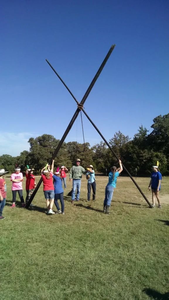 Group outdoors on a grassy field raising two long poles crossed into an X against a blue sky.