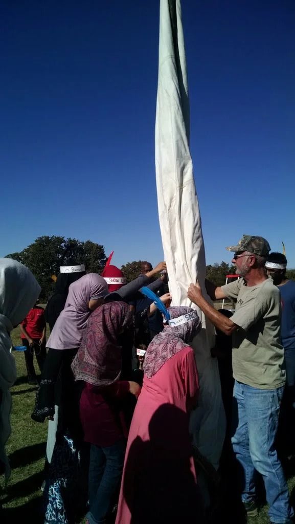 People raising a tall white flag or banner outdoors on a sunny day.