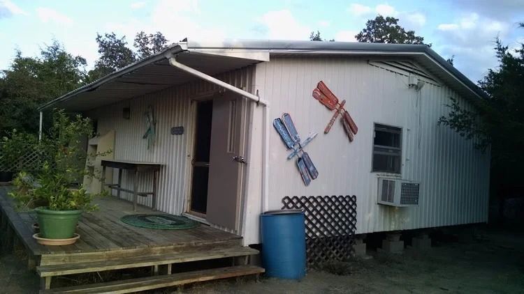 Small beige shed with a porch, colorful airplane decorations, and a blue barrel in a grassy yard
