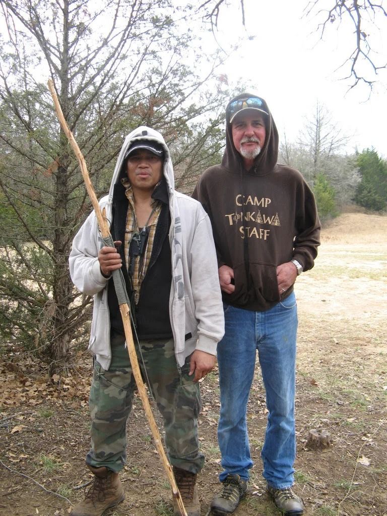 Two hikers standing outdoors, one holding a walking stick, in a wooded area.