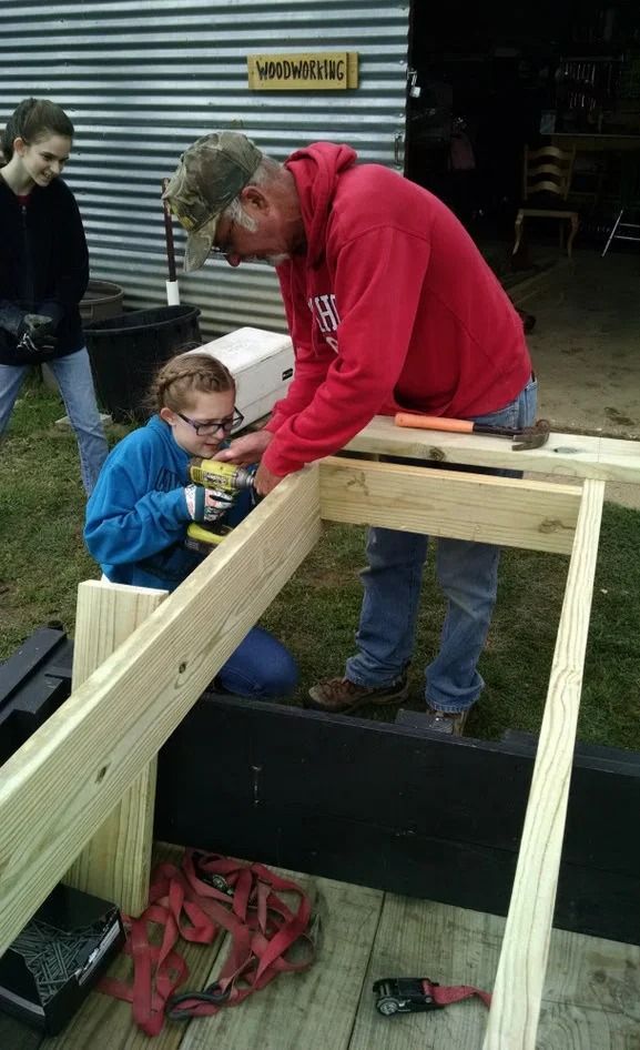 Two people fitting a wooden frame together at a construction site, one in a red hoodie and one in blue.