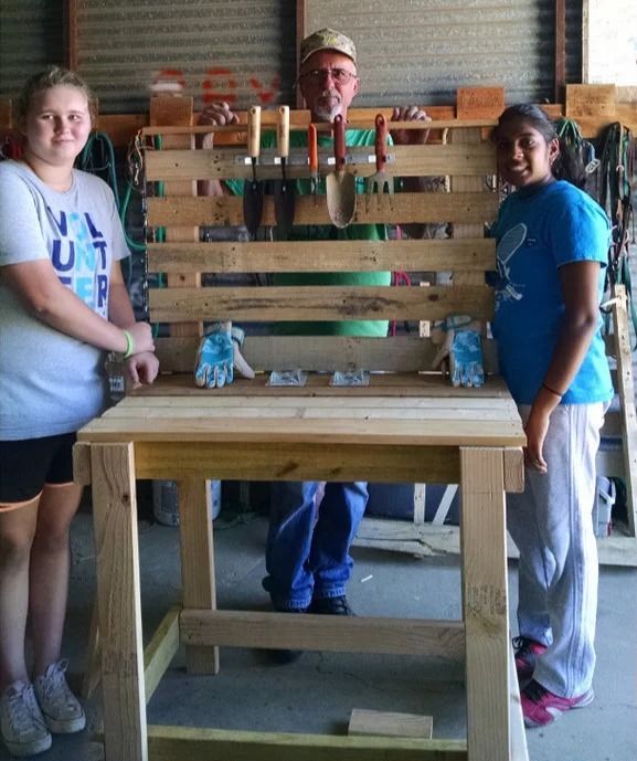 Three people stand behind a wooden table in a workshop, holding a large slatted wooden chair.