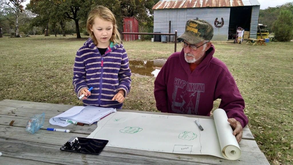Two people sit at a picnic table, writing on paper outdoors near a small shed.