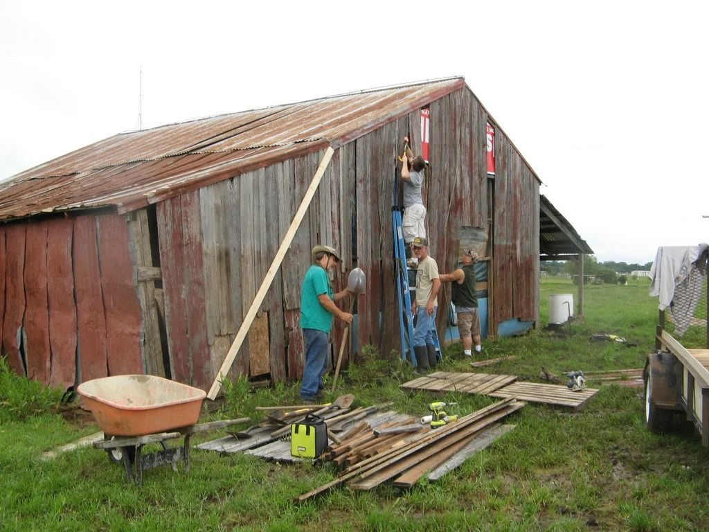 People repairing a weathered red barn with tools and lumber scattered outside