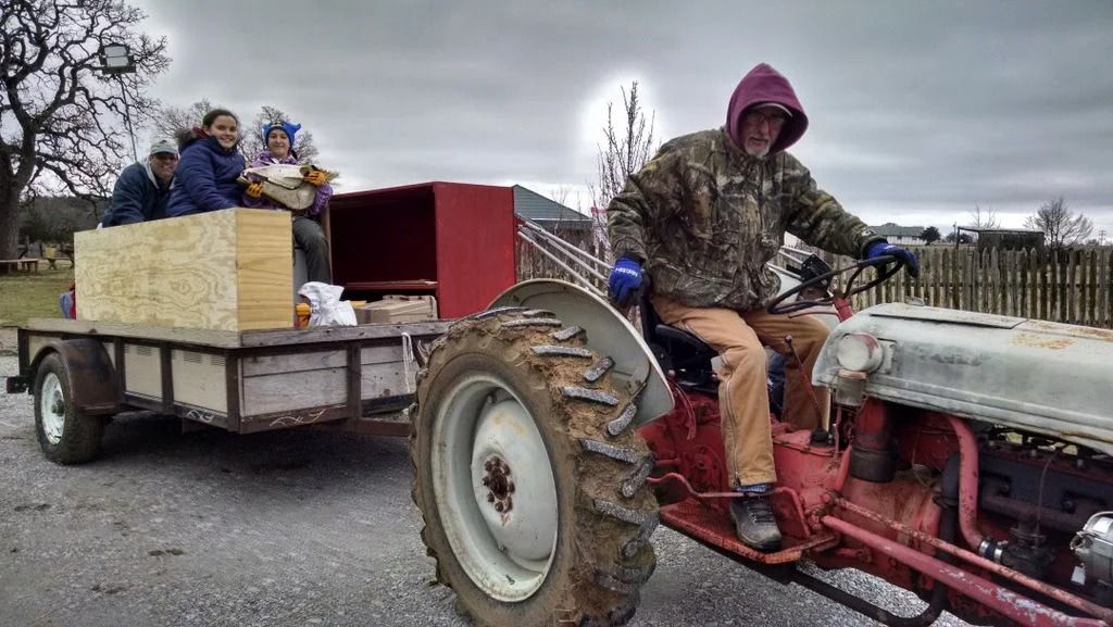 Tractor towing a flatbed trailer with bundled passengers on a gravel road in a rural field
