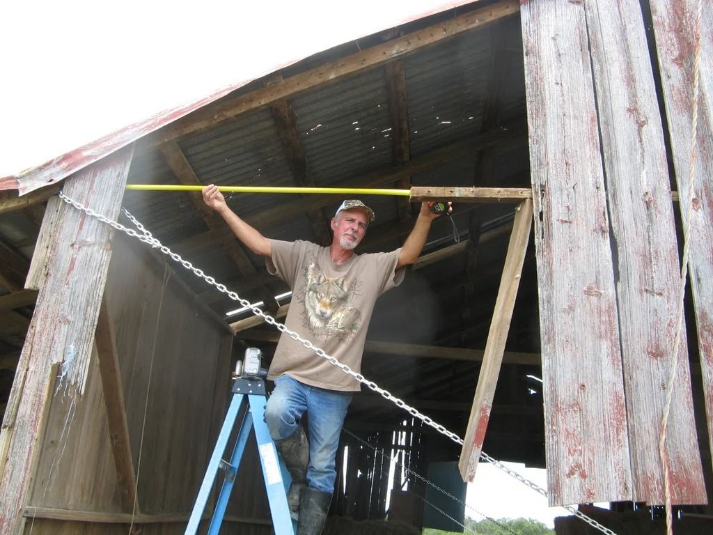 Man measuring a barn opening with a tape measure while standing on a ladder.
