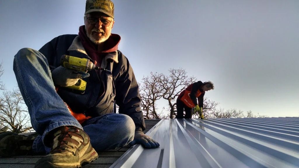 Two workers on a metal roof, one in the foreground crouching with tools and one working in the distance.
