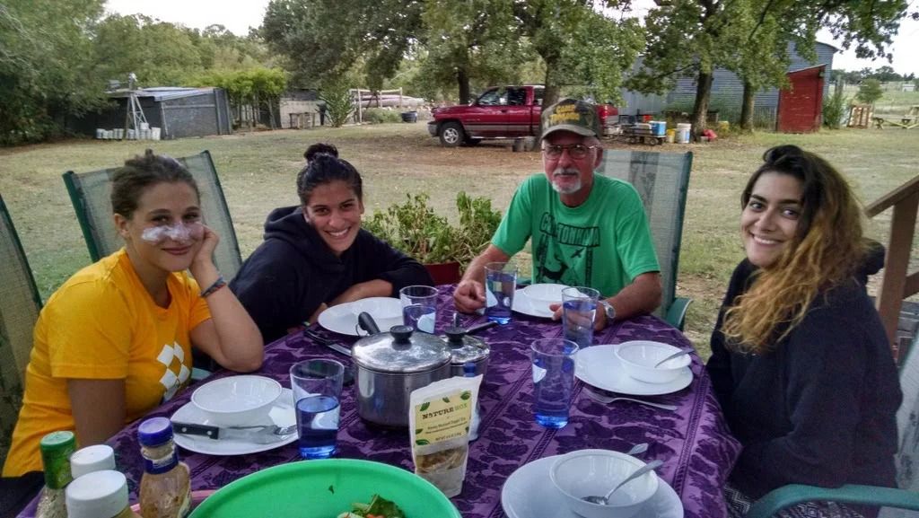 Four people sitting at a picnic table outdoors, sharing a meal under trees.