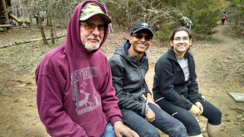 Three people sitting on a wooden bench on a wooded trail, smiling at the camera.