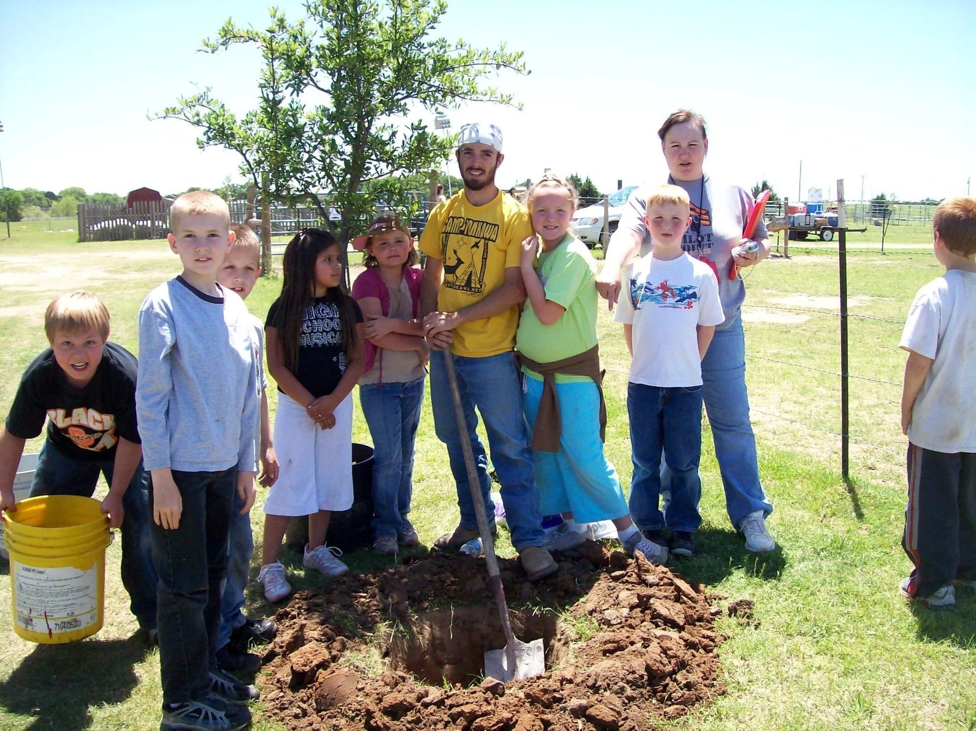 Children and adults stand around a newly planted tree in a sunny park, one person holding a shovel.