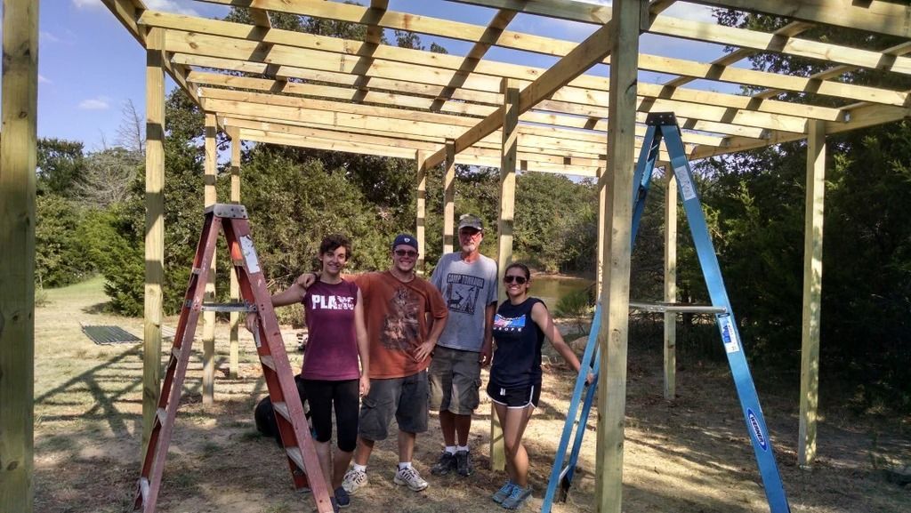 Four people standing under a wooden pergola frame in a grassy outdoor area, with ladders supporting the structure.