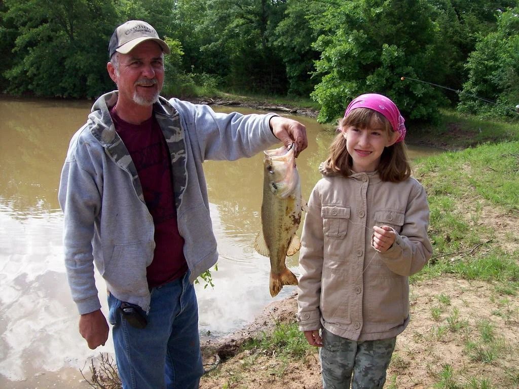 Man holding a large fish beside a smiling child outdoors near a pond
