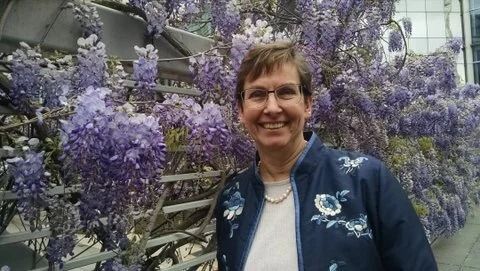 Smiling person standing among purple flowers and a wooden trellis outdoors, wearing a blue jacket.