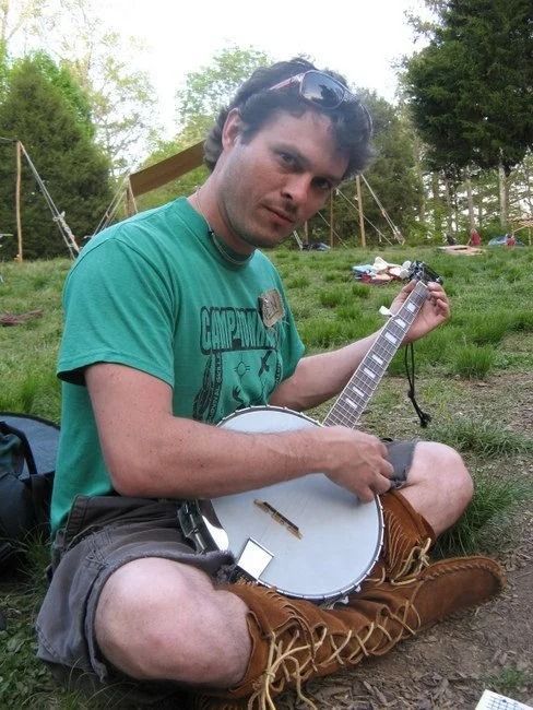 Man playing a banjo while seated outdoors on grass, wearing a green T-shirt and shorts.
