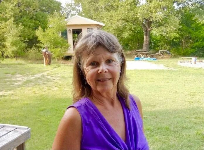 Smiling woman in a purple top standing on a grassy lawn with trees and a small gazebo behind her