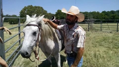 Cowboy in a white hat stands beside a white horse in a grassy fenced field.