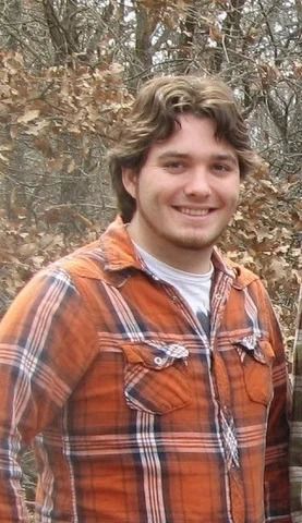 Smiling person in an orange plaid shirt standing outdoors among leafless trees