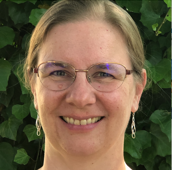 Close-up portrait of a smiling person wearing glasses and earrings, with green leaves in the background