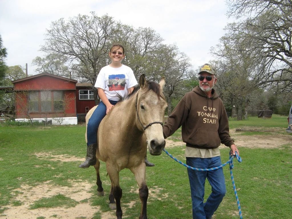 A man leading a tan horse with a woman riding it in a grassy yard near a small house.