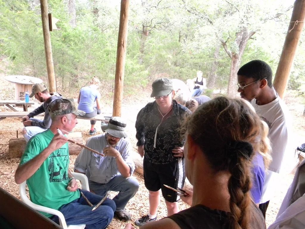 People gathered at a picnic table under trees, talking and sitting outdoors.