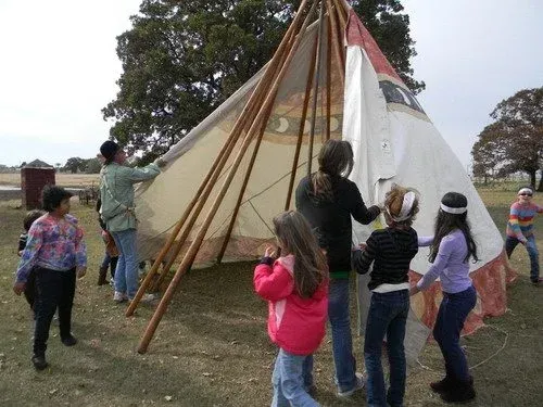 People assembling a large white teepee outdoors, with wooden poles and children standing nearby