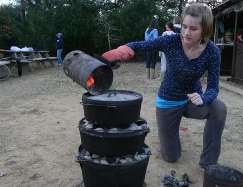 Person in blue sweater pours glowing coals from a barrel into a stacked metal stove outdoors.