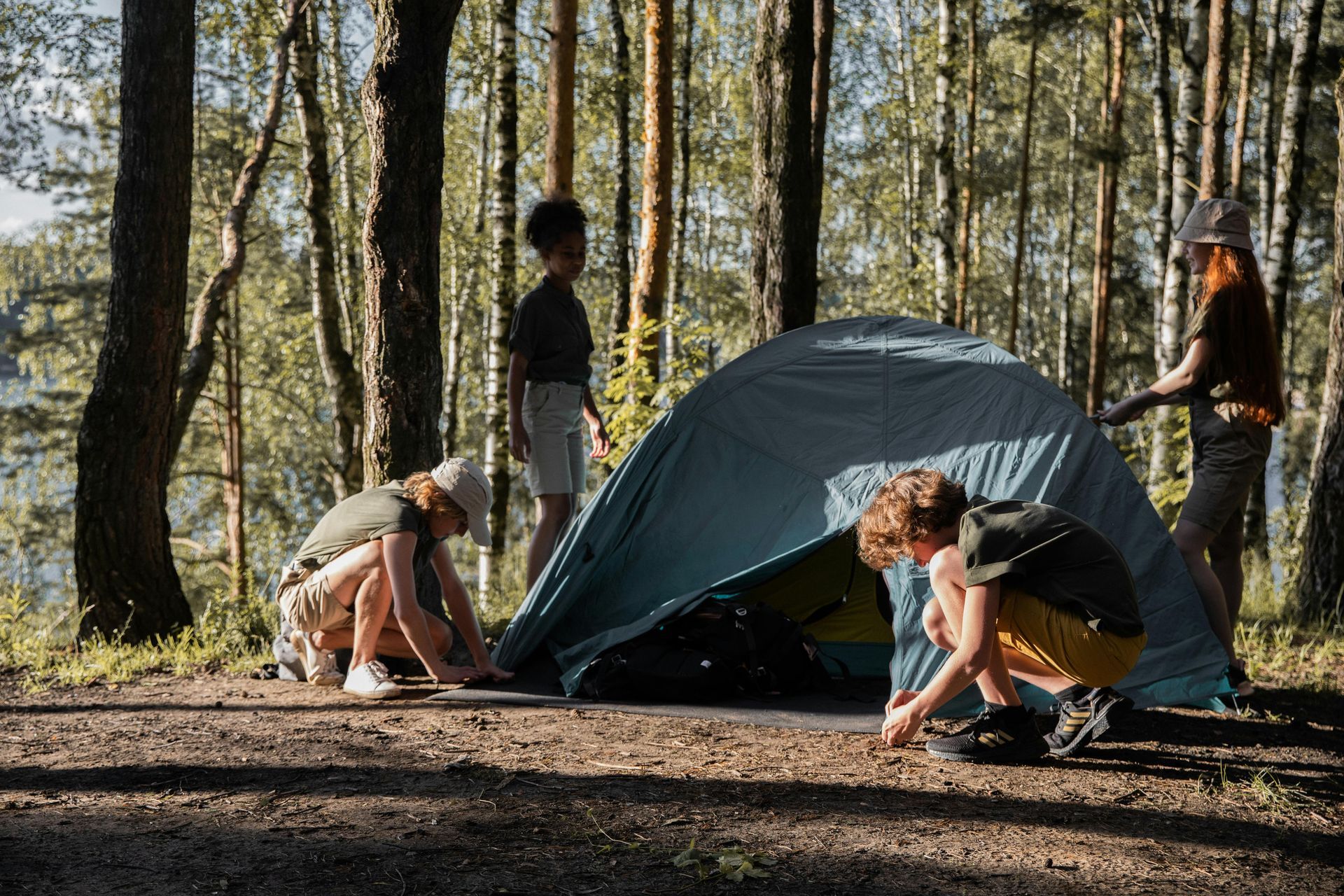 Four people set up a blue camping tent in a sunny, forested area with tall trees.