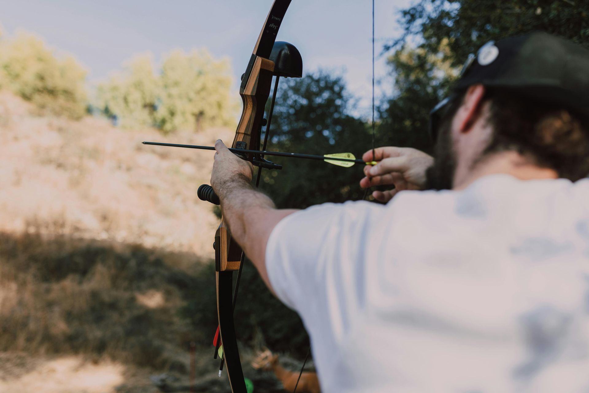 A person wearing a hat and white shirt draws back the string of a compound bow in an outdoor, wooded setting.