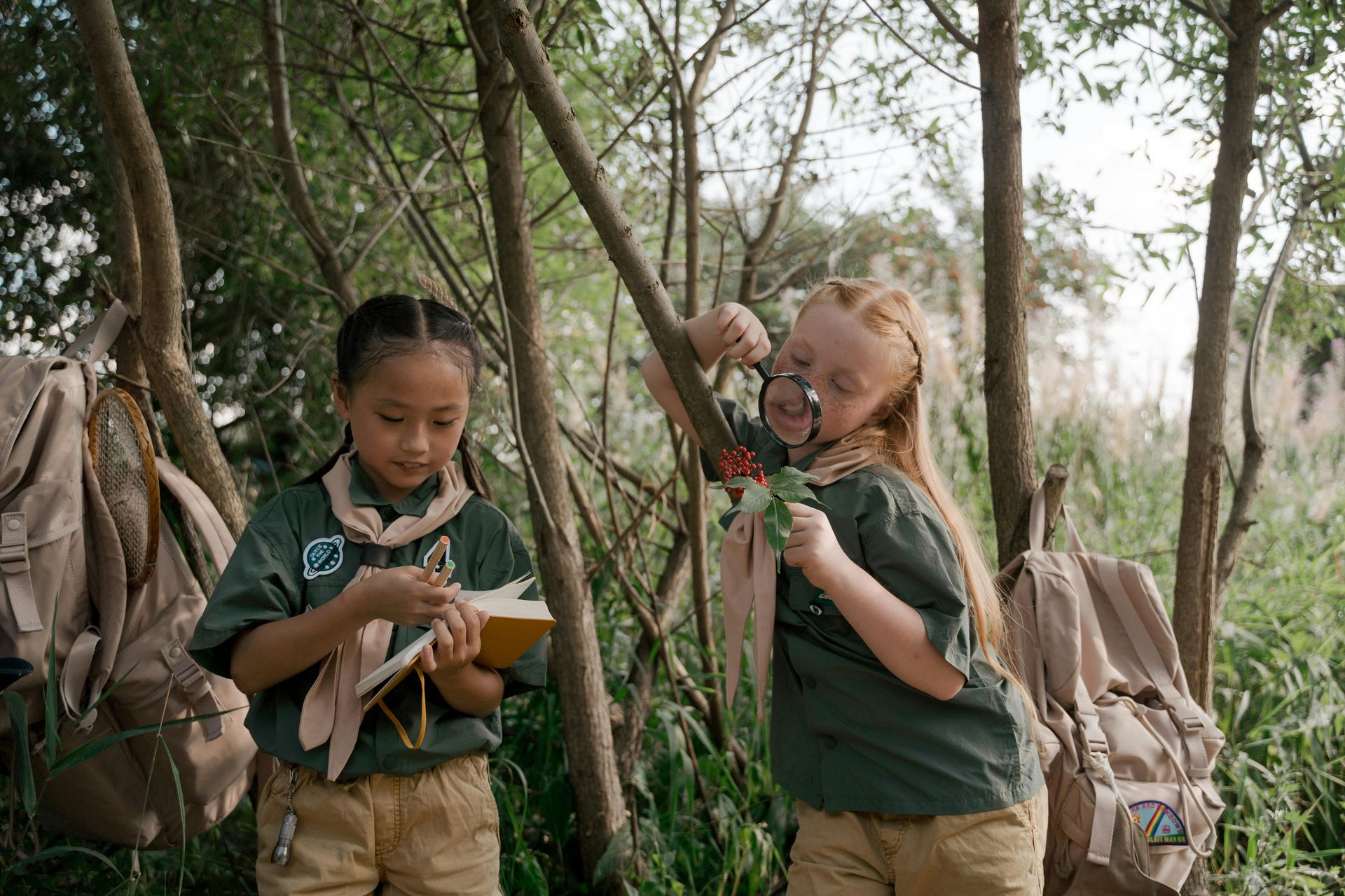 Two young scouts in green uniforms explore a forest, one reading a field guide and the other examining leaves with a lens.