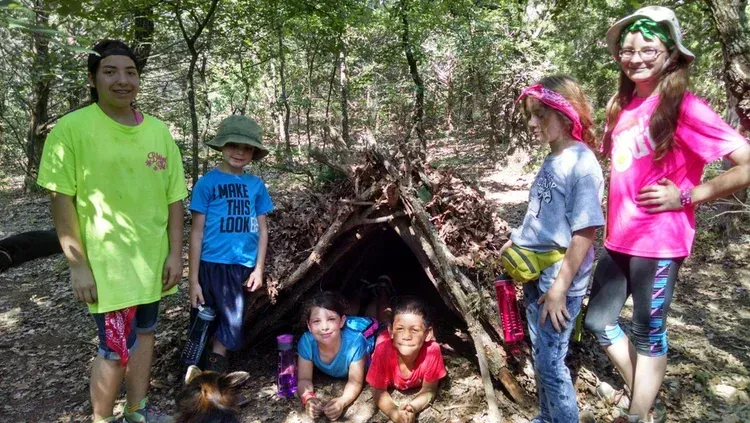 Five children pose beside a small stick shelter in a wooded area, with two crouching inside the opening.