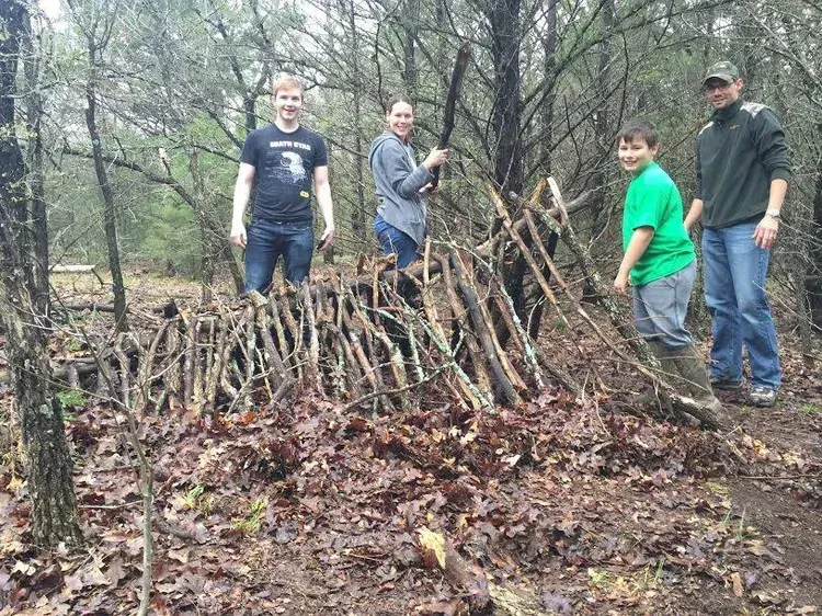 Four people stand beside a large stick-and-branch shelter in a wooded area.