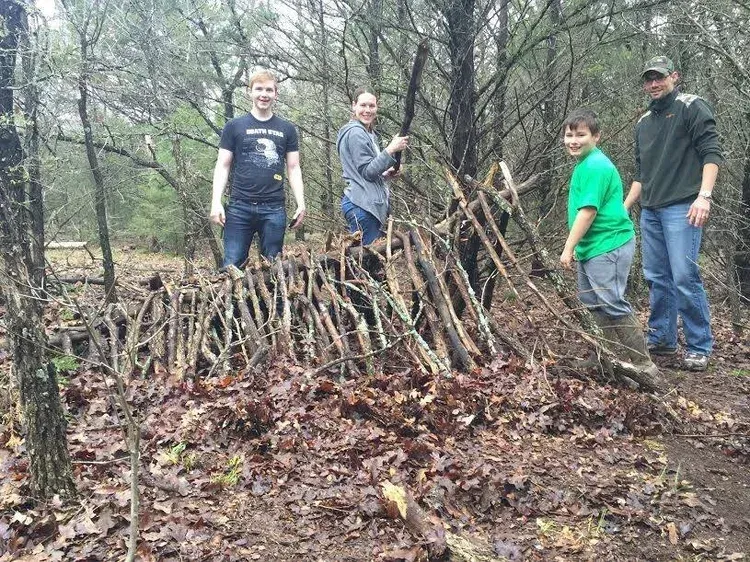 Four people stand by a large brush pile in a wooded area, one holding branches.
