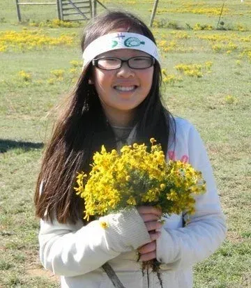 Smiling person outdoors holding a bouquet of yellow flowers, wearing a white headband and glasses.