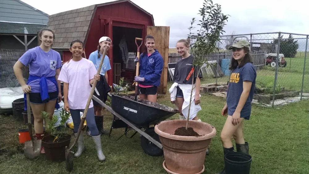 Group of people with garden tools beside a red shed, wheelbarrow, and potted plant in a yard.