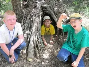 Three children crouch at a twig shelter entrance in a wooded area; one raises a fist and smiles.