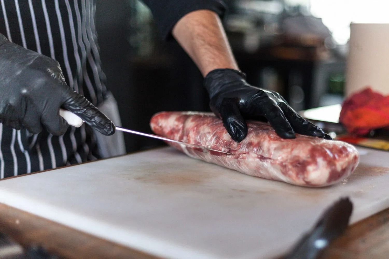 Gloved hands slicing a raw cut of meat on a cutting board in a kitchen