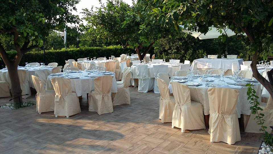 Tavoli per ricevimenti all'aperto apparecchiati per una festa sotto gli alberi; tovaglie e coprisedie bianchi.