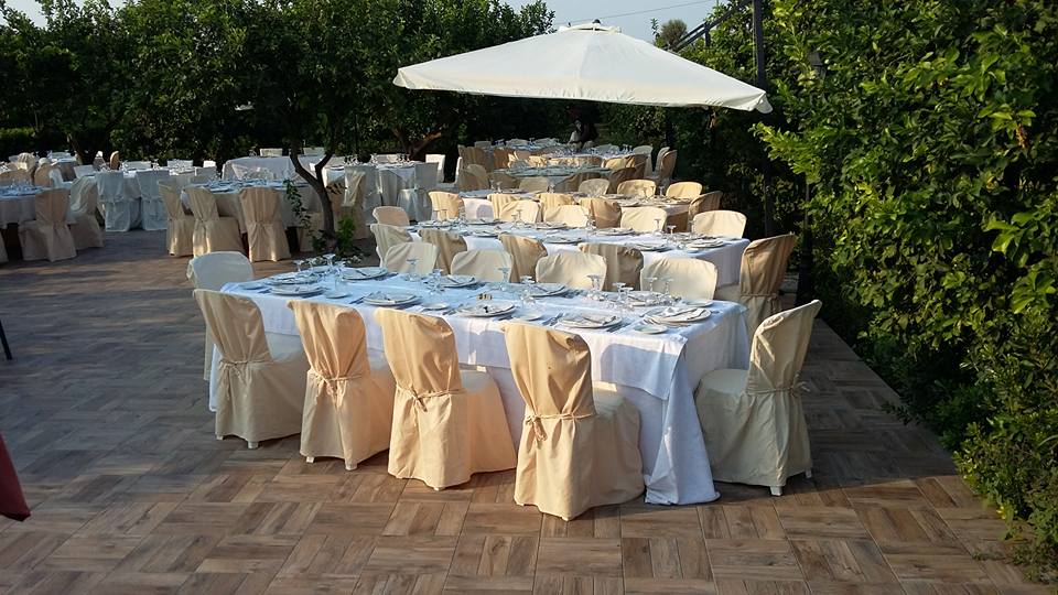 Tavoli per eventi all'aperto apparecchiati con tovaglie bianche, sedie beige e un ombrellone, circondati da alberi.