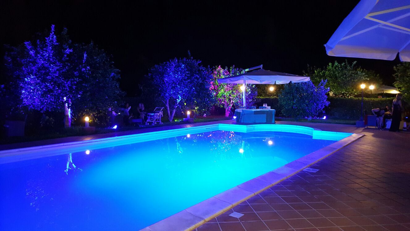 Scena notturna: una piscina si illumina di turchese. Gli alberi e il patio sono illuminati da una luce blu.