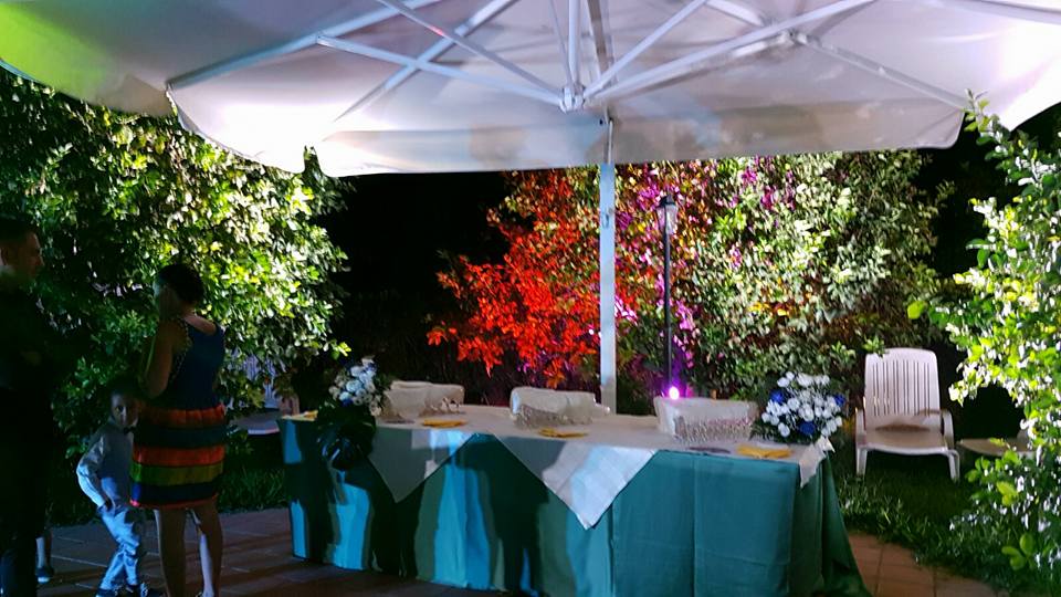 Evento all'aperto, tavoli con decorazioni floreali sotto grandi ombrelloni, circondati dal fogliame, persone che si mescolano.